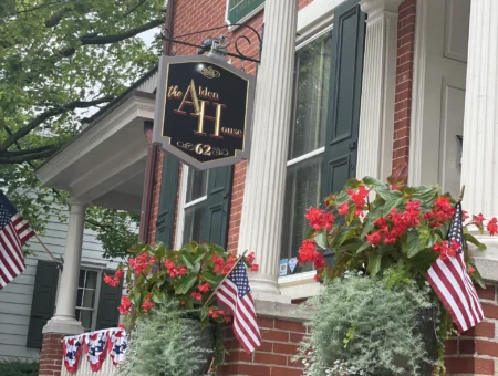 Front entrance of the Alden House Bed and Breakfast with red brick facade, hanging sign, and flower-filled planters with American flags