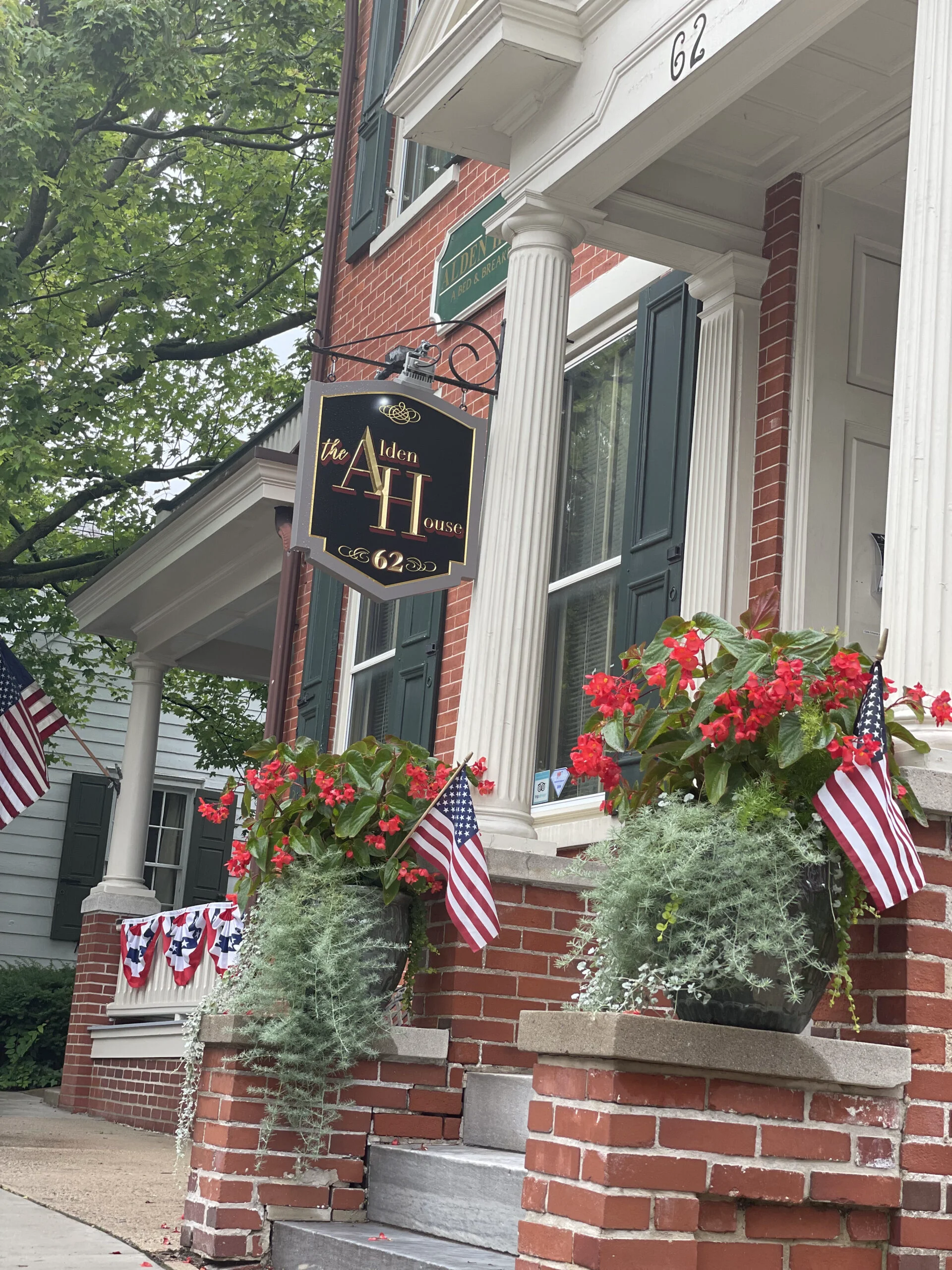 Front entrance of the Alden House Bed and Breakfast with red brick facade, hanging sign, and flower-filled planters with American flags