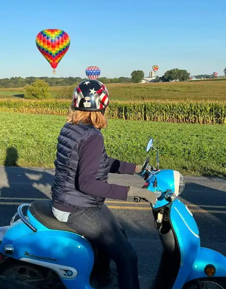 Woman riding a scooter through Amish farmland with hot air balloons in the sky in Strasburg Lancaster County PA