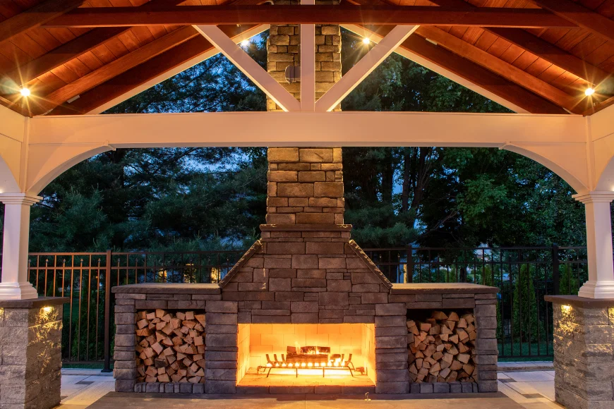 Clymer's Landscaping lit outdoor stone fireplace with cordwood under cedar timber pavilion at dusk in Lancaster PA