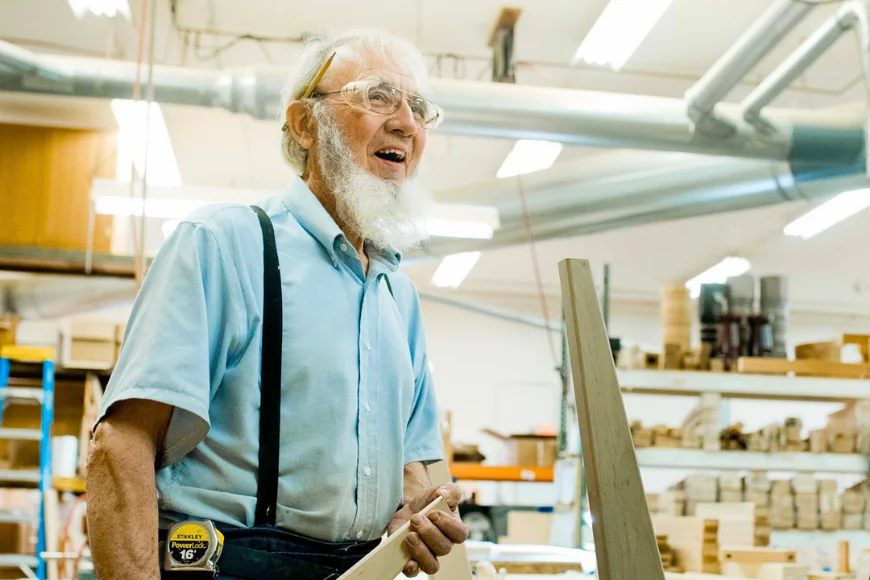 Elderly woodworker with white beard holding lumber in a furniture workshop