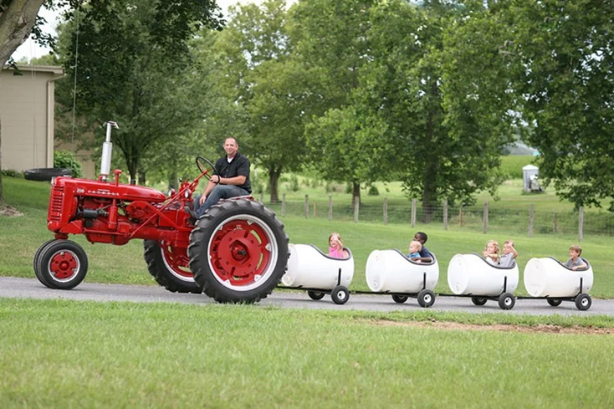 Red antique tractor pulling a barrel train of kids at Landis Farm, with a man driving and passing rolling pastureland in the background