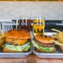 Two burgers with fries and draft beers on metal trays at a restaurant in Lititz, PA, with outdoor seating visible in the background