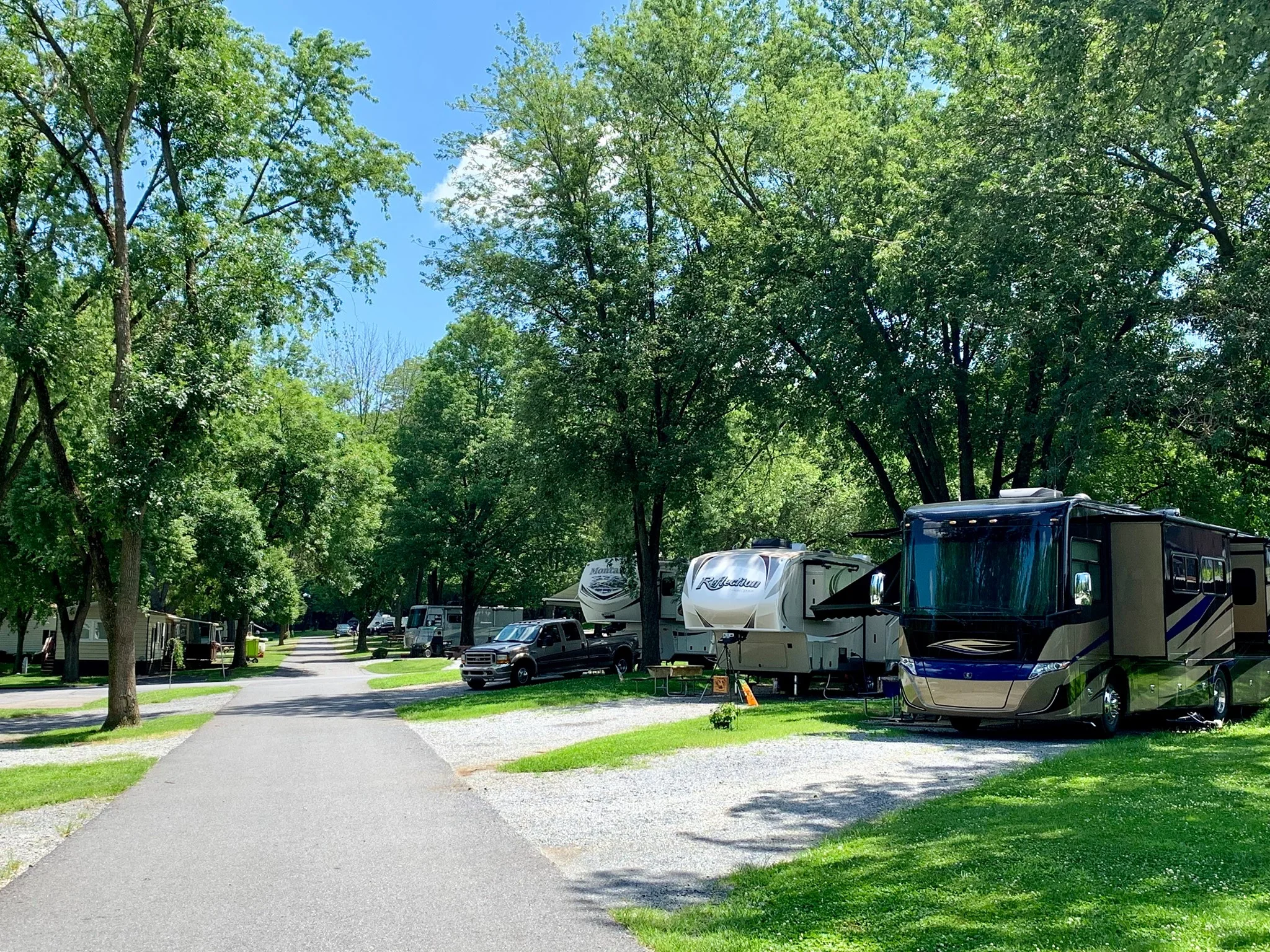 Old Mill Stream Campground gallery image: paved campground road lined with RVs, campers, and a motorhome parked among tall shade trees in Lancaster, PA