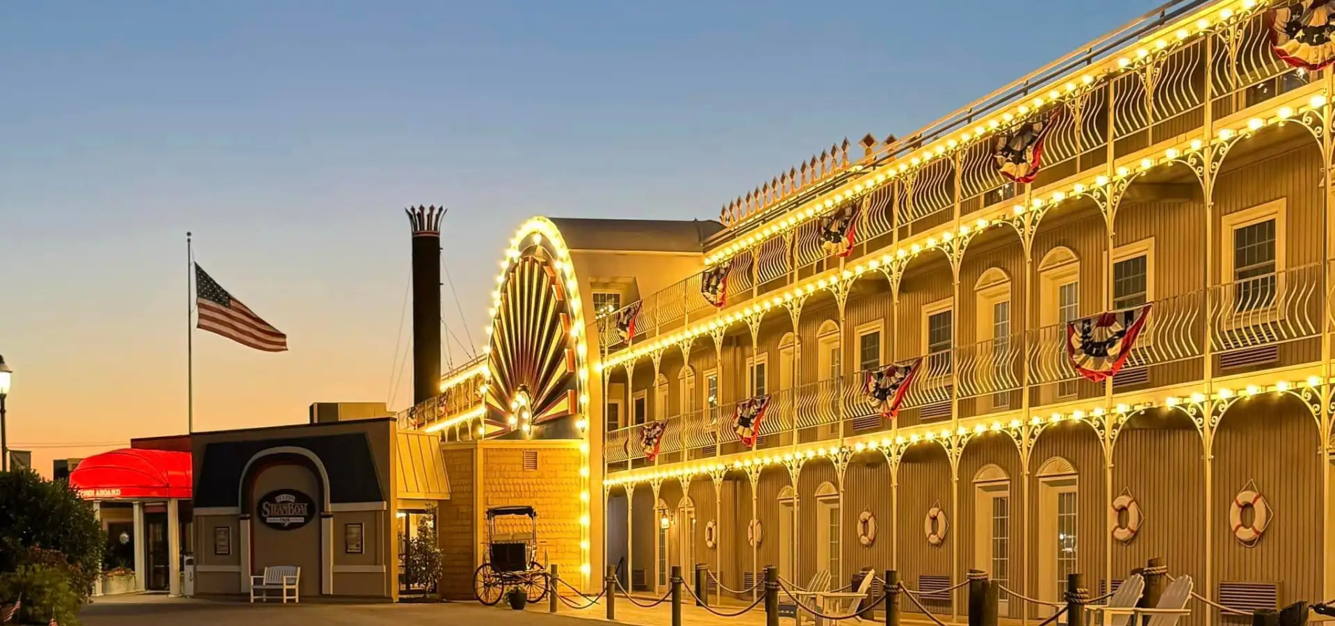 Steamboat-themed hotel exterior on Route 30 East, with wraparound decorative railings, string lights, patriotic bunting, and a large steamboat wheel accent along the facade