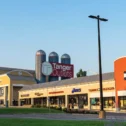 Tanger Outlets storefront row along Route 30 East, featuring a barn-style facade with silos and storefronts for Asics, Tommy Hilfiger, and Kay Jewelers under a clear sky