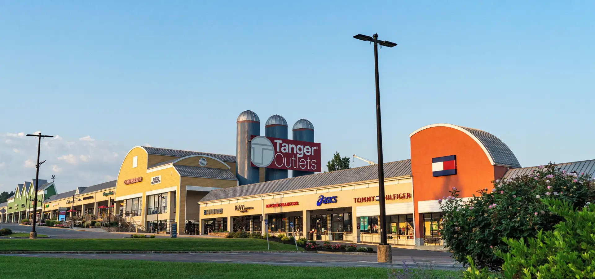 Tanger Outlets storefront row along Route 30 East, featuring a barn-style facade with silos and storefronts for Asics, Tommy Hilfiger, and Kay Jewelers under a clear sky