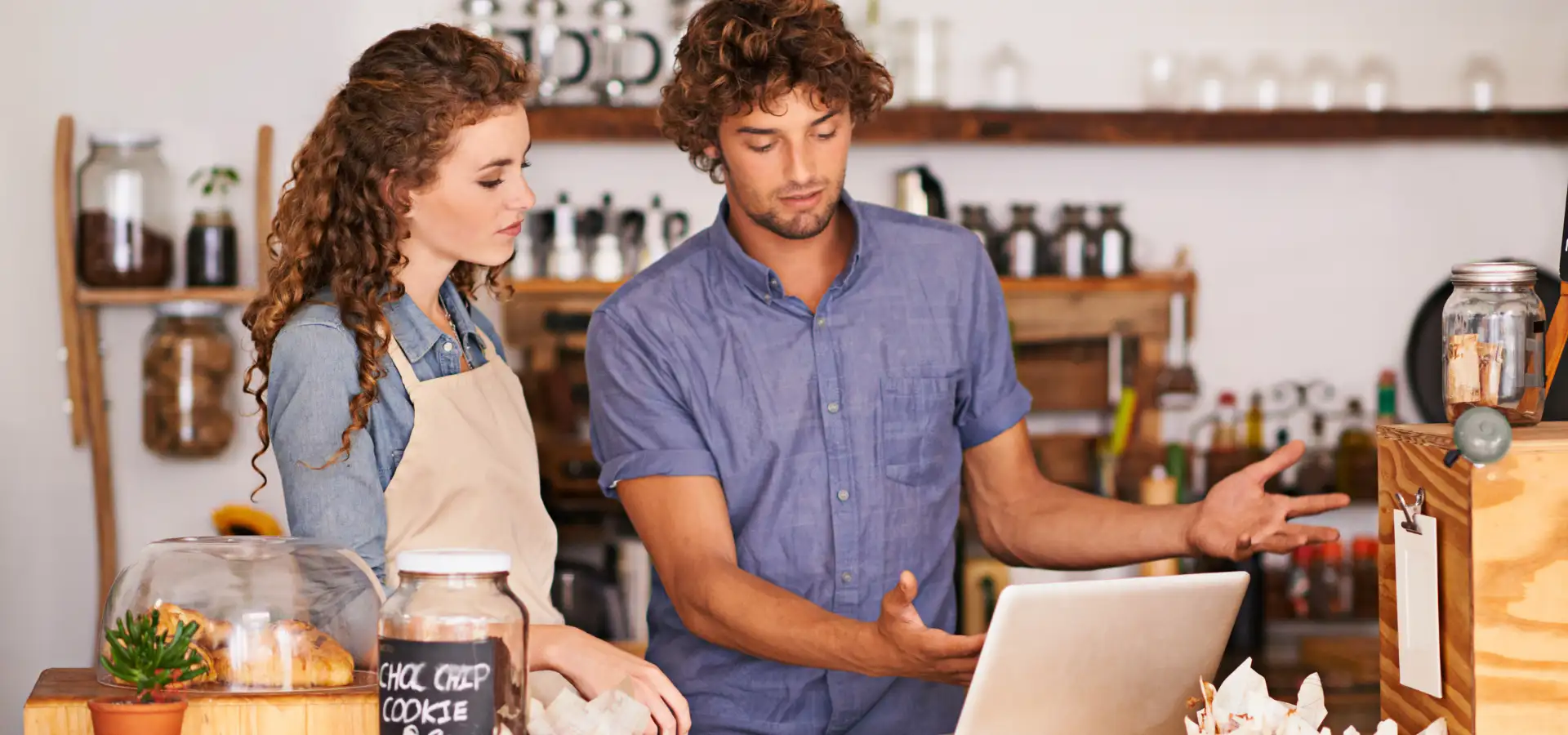 Small Business Saturday scene at a shop in Lancaster, PA, with an owner and employee reviewing a laptop near jars of coffee and baked goods on the counter