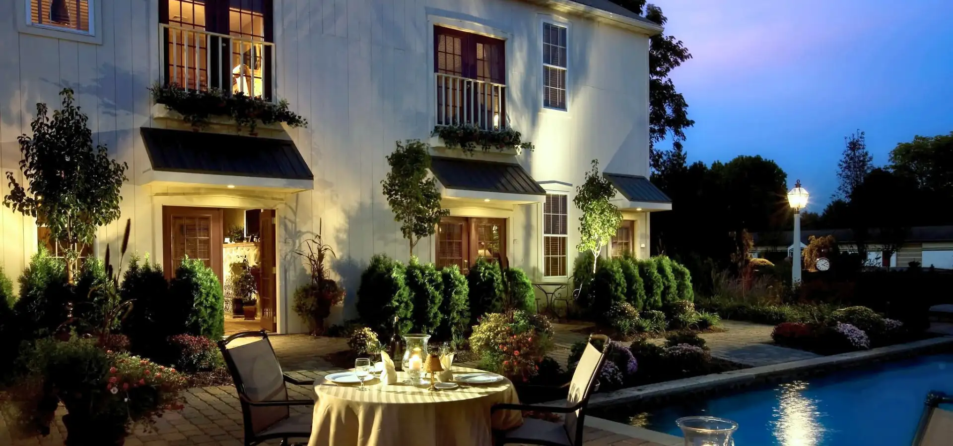 Poolside patio dining setup at Inn at Leola Village near suburban Lancaster, PA, with a set table, string lights, illuminated pool, and lush landscaping at dusk