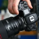 Person's hand holding a Canon EOS R5 camera with an 85mm lens, photographed at a shop in suburban Lancaster, PA, with denim fabric visible in the background
